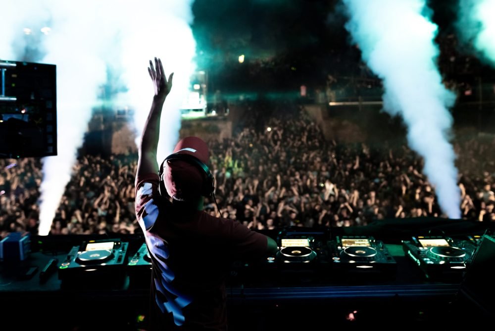Rear view of a DJ playing music in front of excited crowd at music festival by night.